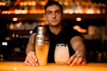 close-up front view on shaker and glass with ice and hands of bartender on bar counter
