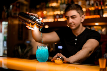 selective focus on glass with iced blue cocktail on bar and bartender pouring liquid into it from shaker