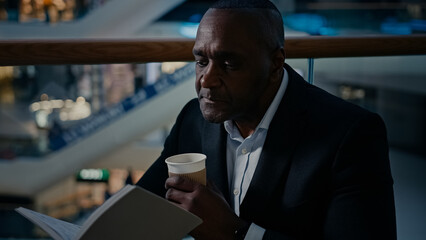 Ethnic male African man businessman employer entrepreneur drinking tea aromatic hot coffee at table in cafe cafeteria restaurant drink latte cappuccino enjoying reading book read notebook text study