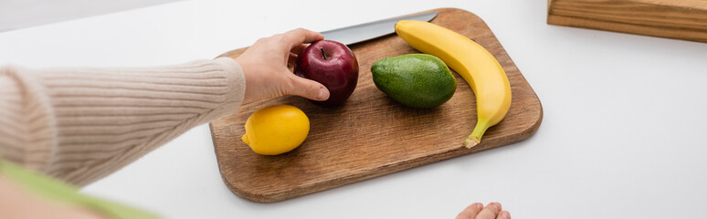 Cropped view of woman holding apple near fruits on chopping board at home, banner