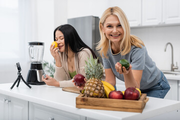 Smiling woman holding fruit near asian friend and smartphone on tripod in kitchen