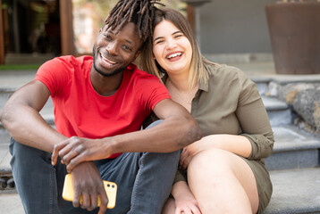 A smiling multi-ethnic couple sitting outside looking at the camera smiling