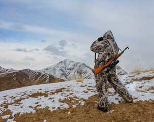Huntsman in winter camouflage with an old carbine inspects the mountains through binoculars.