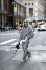 full length of blonde woman in knitted sweater and sunglasses walking with hand in pocket on urban street in New York