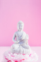 Decorative white Buddha statuette sitting on the peony flower pillow altar on the pink background. Meditation and relaxation ritual. Buddha birthday. Minimalism.Vertical card. Selective focus.