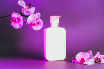 Obraz na płótnie Canvas Pink plastic bottle with mock up space on purple neon background with orchid flowers. Natural cosmetic mockup. Selective focus. Copy space.