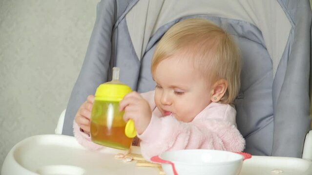 A cute one-year-old girl drinks compote from a bottle with a pacifier, from a sippy cup, eats cookies sitting in a high chair