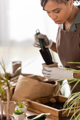 african american florist holding garden scoop and flowerpot near wooden box with soil and blurred plants