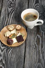 A cup of black coffee in a white cup. Chocolate in foil and chocolates. On pine boards.