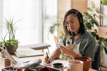 young african american florist in eyeglasses looking at smartphone and writing order in notebook in flower shop