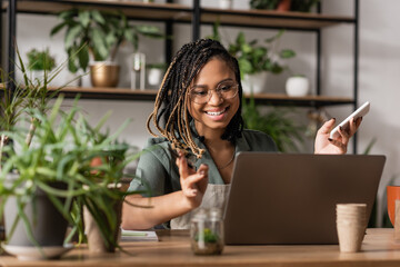 happy african american florist holding smartphone and gesturing during video call on laptop near blurred plants - obrazy, fototapety, plakaty