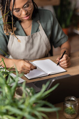 Fototapeta na wymiar african american florist in apron writing order in blank notebook near green plants on blurred foreground