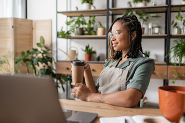 happy african american florist in eyeglasses and apron sitting with paper cup near blurred laptop in flower shop