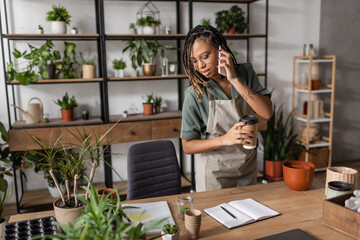 african american florist with paper cup talking on mobile phone and looking at blank notebook near potted plants