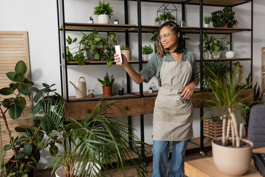 smiling african american florist in apron holding cellphone while standing with rack of potted plants during online order