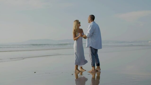 Beautiful young couple dancing at seashore, spending time together on holiday at sunset. Caucasian man in blue shirt twirling attractive blonde woman in sunglasses under his arm. Holiday, love concept