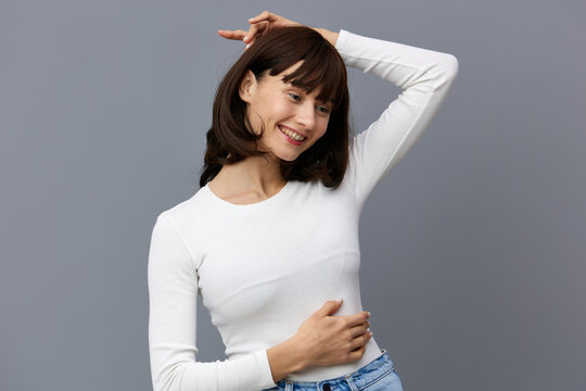 a beautiful woman stands full-face against a dark background in a tight white T-shirt, gently straightens her disheveled hair with her hand, putting her other hand on her stomach