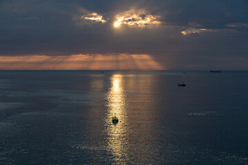 A lone boat sits on a calm sea, as crepuscular sun rays break through from behind the clouds at sunrise