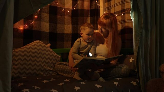 A young pregnant mother and a little son in pajamas in a bed house shine a flashlight into a book and discuss what they saw. In the background is a Christmas garland. High quality 4k footage