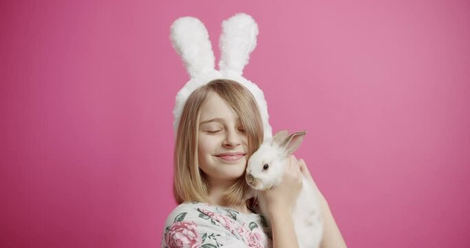 Happy girl with cute bunny. Static shot of glad blond girl with bunny ears smiling and touching noses with adorable white rabbit during Easter celebration against pink background. Happy Easter