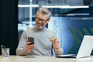 Cheerful and smiling gray-haired businessman inside a modern office makes purchases in an online store, the man holds a bank credit card and a smartphone in his hands, the buyer is satisfied choice.