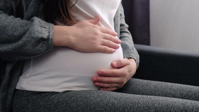 Close up of unknown pregnant woman sitting on comfy couch, puts palms on big belly feel unconditional love to unborn baby. Third trimester of pregnancy, happy maternity, expectant mother concept