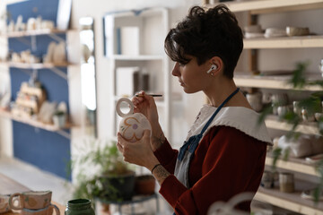 Concentrated Italian woman artist using brush applies paint on handmade dishes made of natural...