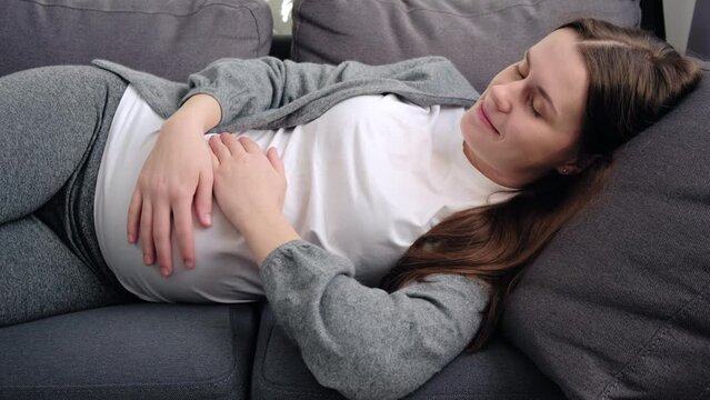 Happy adorable inspired young pregnant female stroking belly, feeling unconditional love to unborn baby, expressing caress and devotion, enjoying carefree serene weekend lying on cozy sofa at home