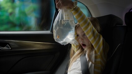 Happy excited Caucasian girl riding on a back seat of a car, bringing home a goldfish in a bag from...