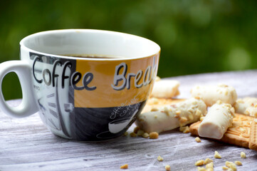 Delicious breakfast: a cup of black tea or coffee and biscuits scattered on a wooden board against the background of a blurred greenery of trees