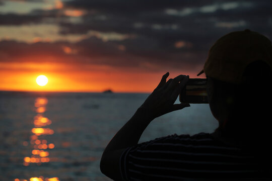 Asian female tourist taking a picture of the ocean sunset with her smartphone camera

