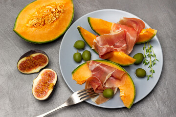 Jamon on slice of melon, green olives and thyme sprigs on gray plate.