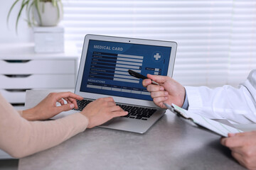Fototapeta premium Doctor and patient using laptop to fill out medical card at table in clinic, closeup