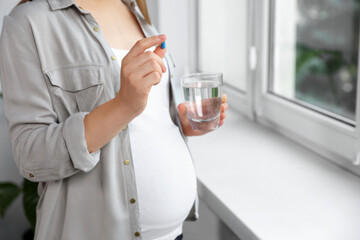 Pregnant woman holding pill and glass of water near window indoors, closeup