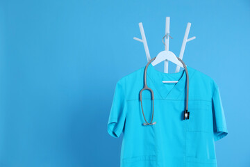 Turquoise medical uniform and stethoscope hanging on rack against light blue background. Space for text