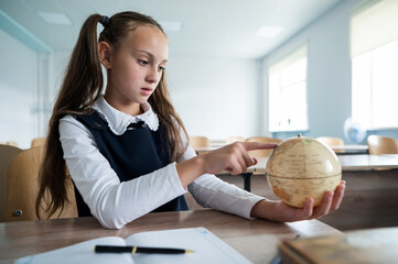 Caucasian schoolgirl sits at her desk at school and studies the globe. 