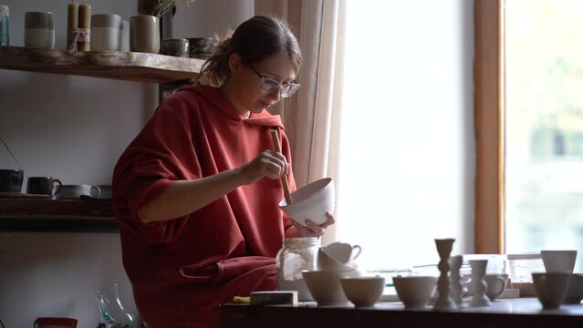 Professional lone woman artist applying protective varnish to crockery restoring antique porcelain service. Concentrated casual girl restorer sits at table in workshop painting plate with white paint