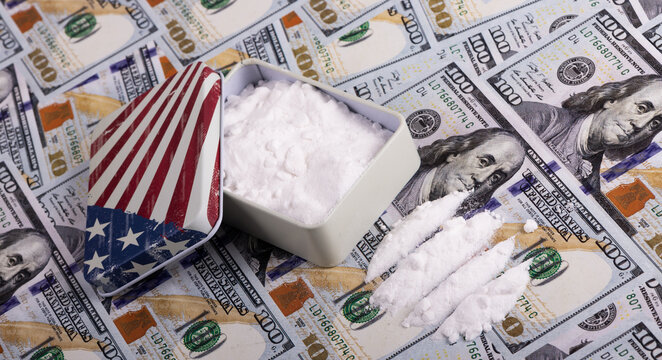 A small metal box with an image of the American flag is filled with white powder, symbolizing drugs, against the backdrop of 100 US dollar bills.