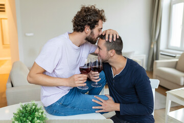 LGBTQ+ couple embracing each other and drinking wine indoors. Two romantic young male lovers looking at each other while sitting together in their living room. Young gay couple being romantic at home.