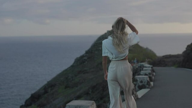 A girl walking towards the lookout to see a landscape of green mountains with the ocean and sunset clouds