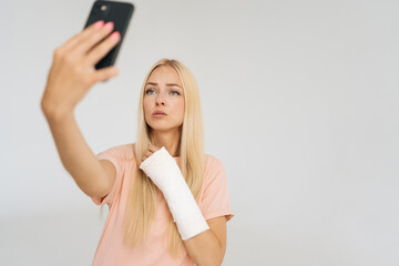 Studio portrait of serious young blonde woman with broken arm wrapped in plaster bandage looking to smartphone screen with puzzled face during video call, standing on white isolated background.