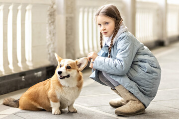 Portrait of a toddler girl in a warm blue coat with a corgi dog in the park. Spring, International Women's Day March 8.