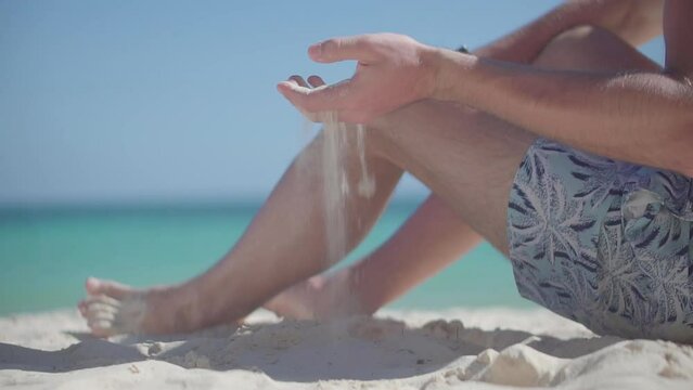 man sits on sandy beach, letting sand slip through fingers
