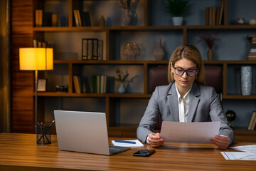 Close up focused young businesswoman working with documents in office, sitting at work table with laptop, serious employee checking financial report, project statistics, reading correspondence