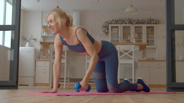 Charming motivated active sporty fit senior woman performing renegade row exercise, toning obliques, rhomboids, and triceps, developing upper body strength while working out at home.