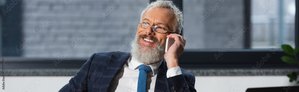 Wall mural smiling businessman in formal wear talking on smartphone in office, banner