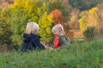 Naklejka na ściany i meble Little siblings resting in nature. Blond boy and girl sit on grass and eat apples. Children on picnic on autumn day.