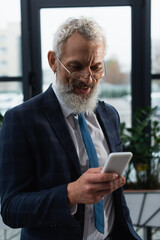 Positive businessman in suit using mobile phone in office