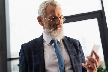 Mature bearded businessman in eyeglasses using blurred smartphone in office