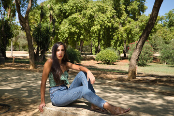 Young woman, brunette, slender, dressed in green T-shirt and jeans, sitting on a stone bench, alone, sad, serious. Concept loneliness, sorrow, sadness.
